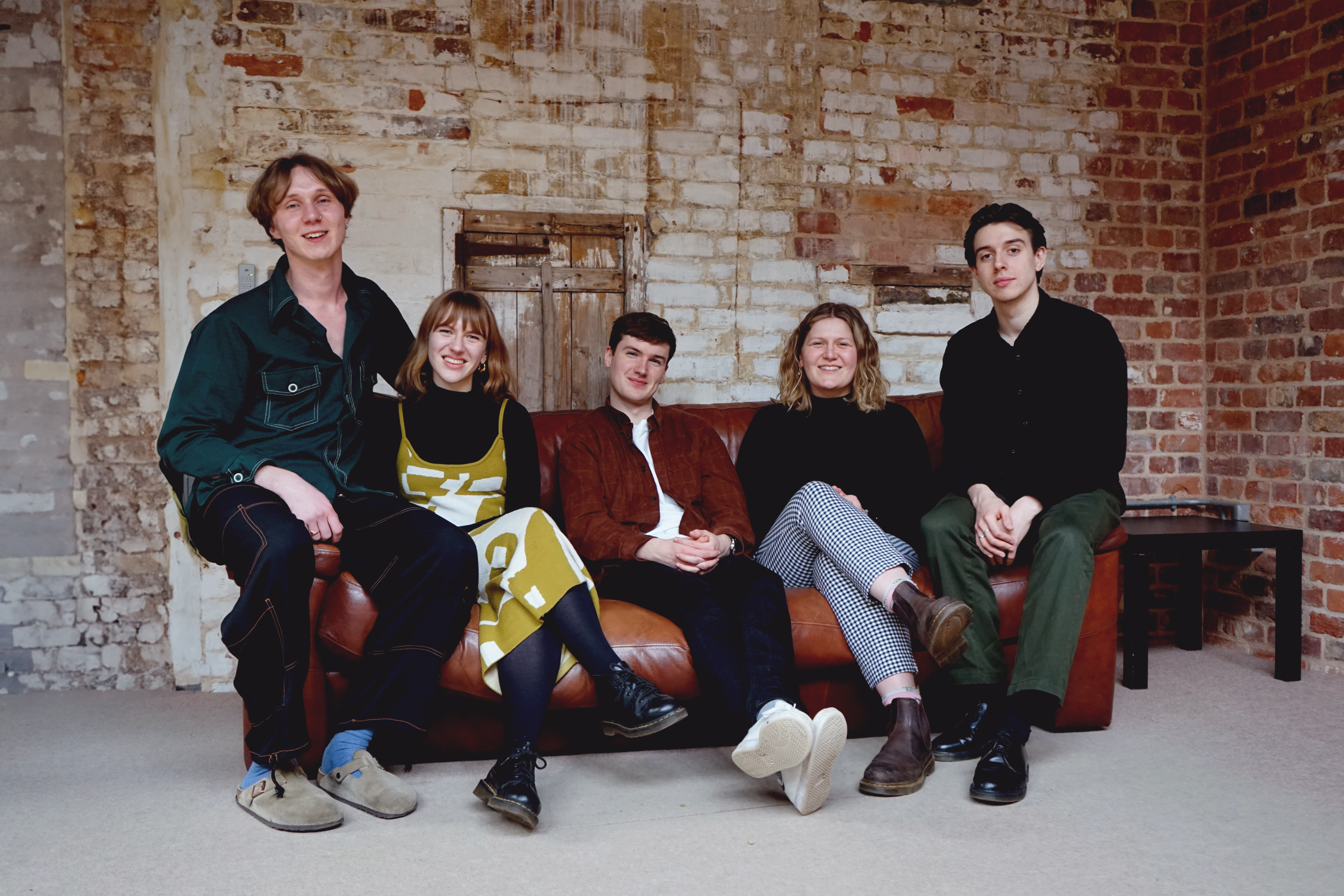 A row of five people sit on a sofa in an empty space with a brick wall behind them.