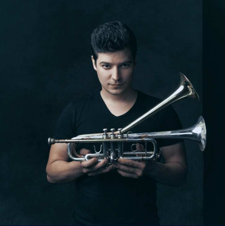 Tamas holds a trumpet in front of a black backdrop.