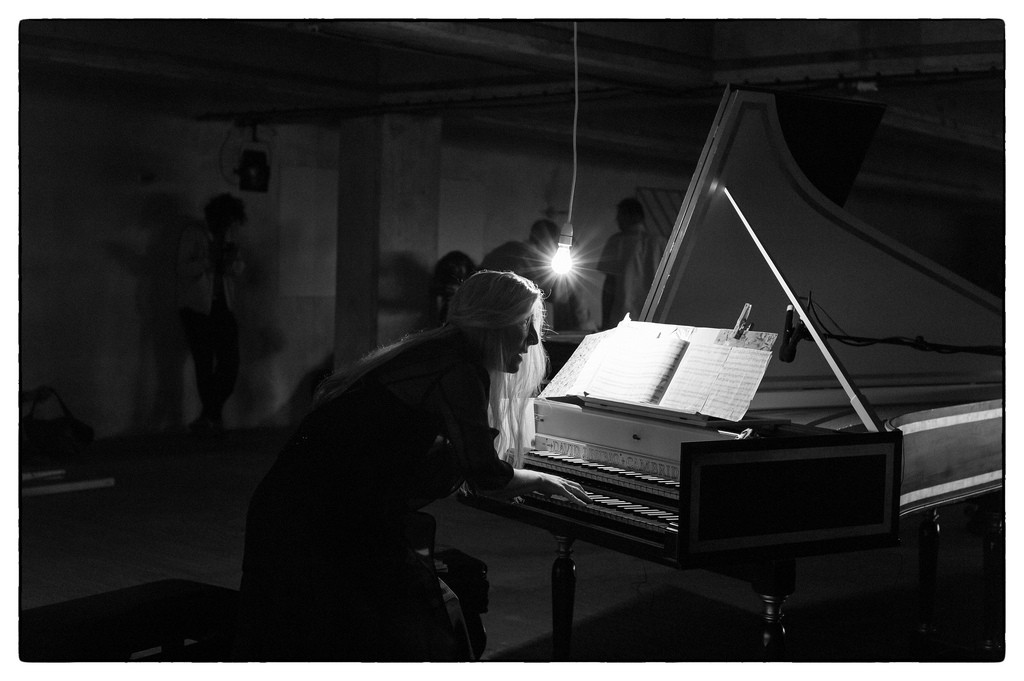 B/w image of Jane Chapman perched in front of a harpsichord over which a lightbulb is dangling