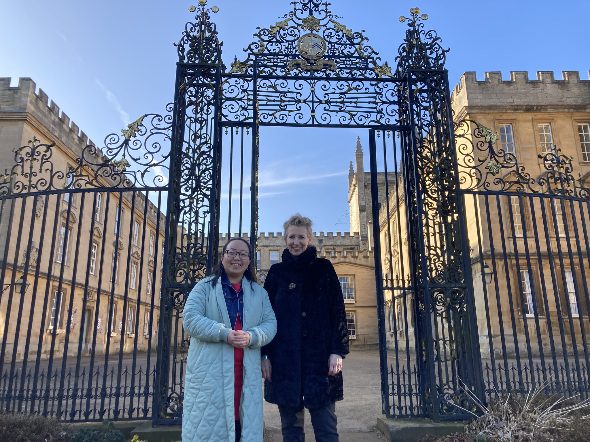 Two women smile at the camera, with an elaborate iron screen and Georgian quad in the background