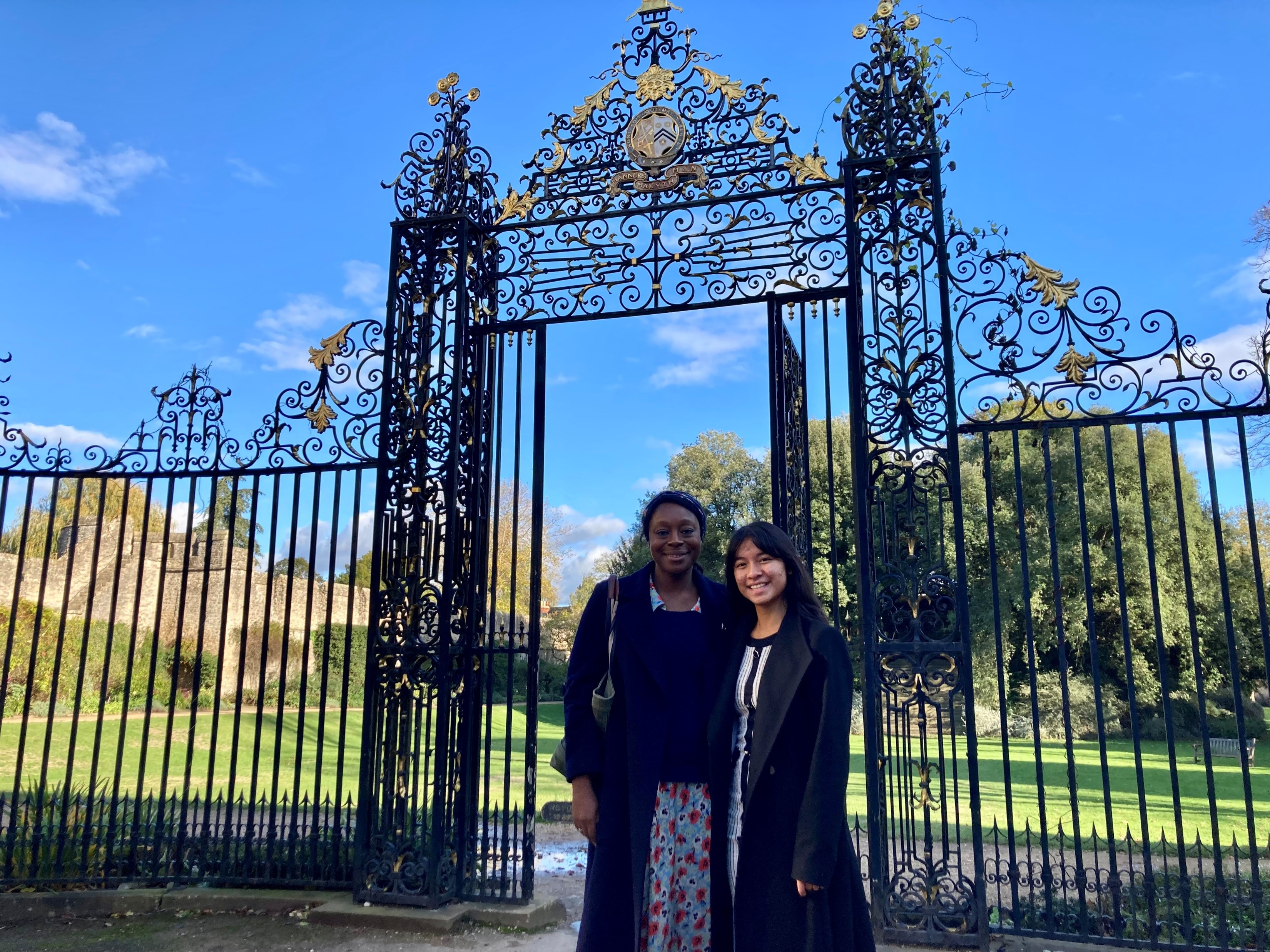 Grace and Ei standing in front of an elaborate iron screen in Garden Quad, with city wall behind