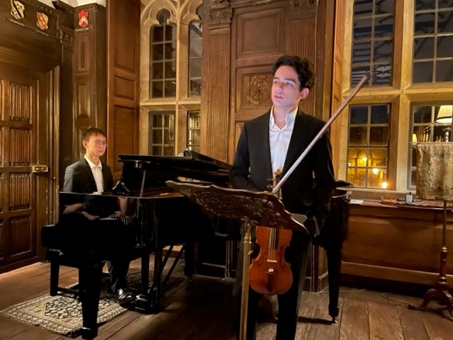 Student holding a violin while another sits behind at a piano