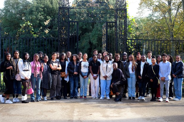 A large group of Black British students smiling at the camera while standing in front of an iron screen and decorative garden mound