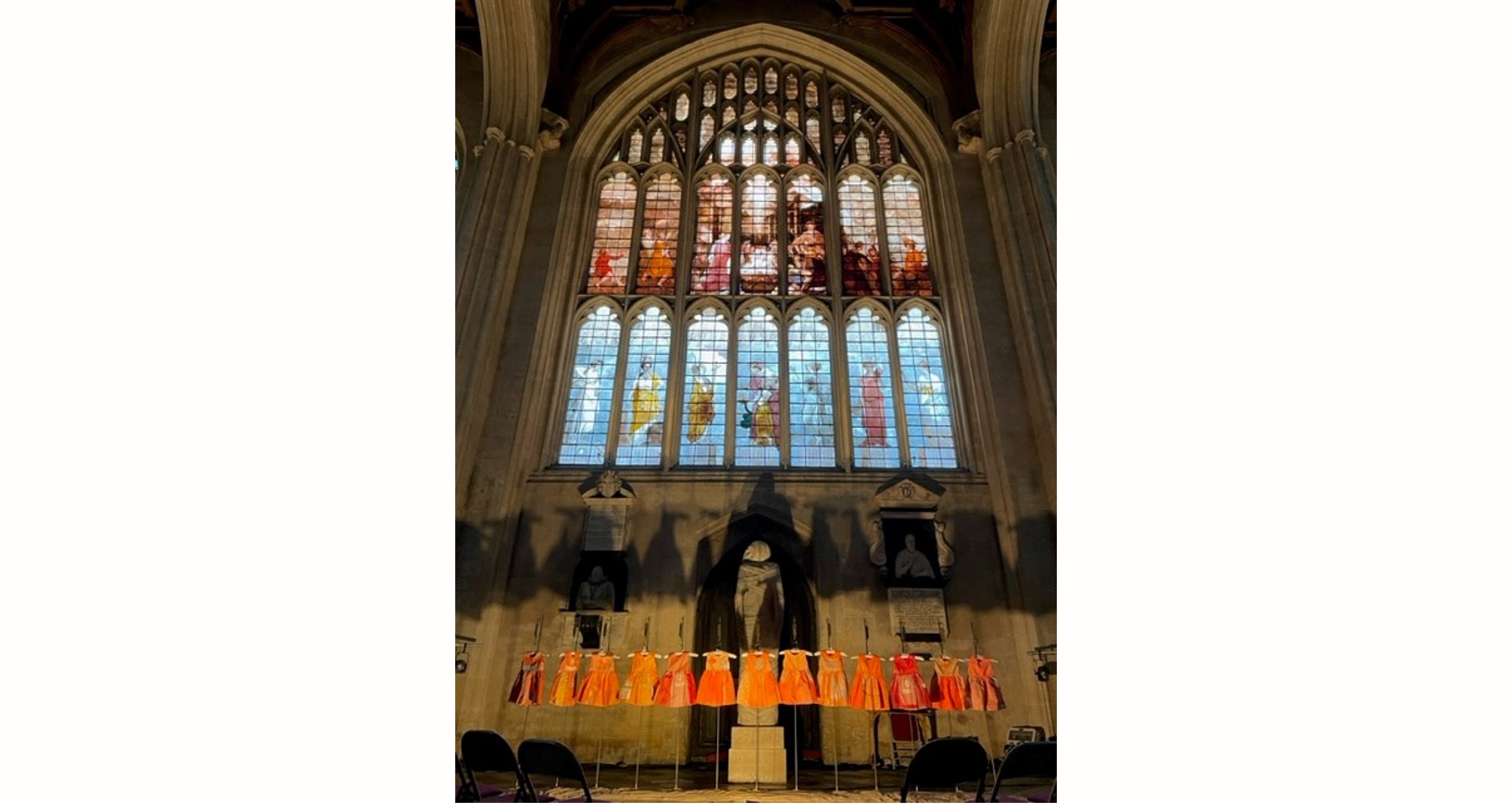 Thirteen orange dresses lined up in a row underneath an ornate stained glass, arched window