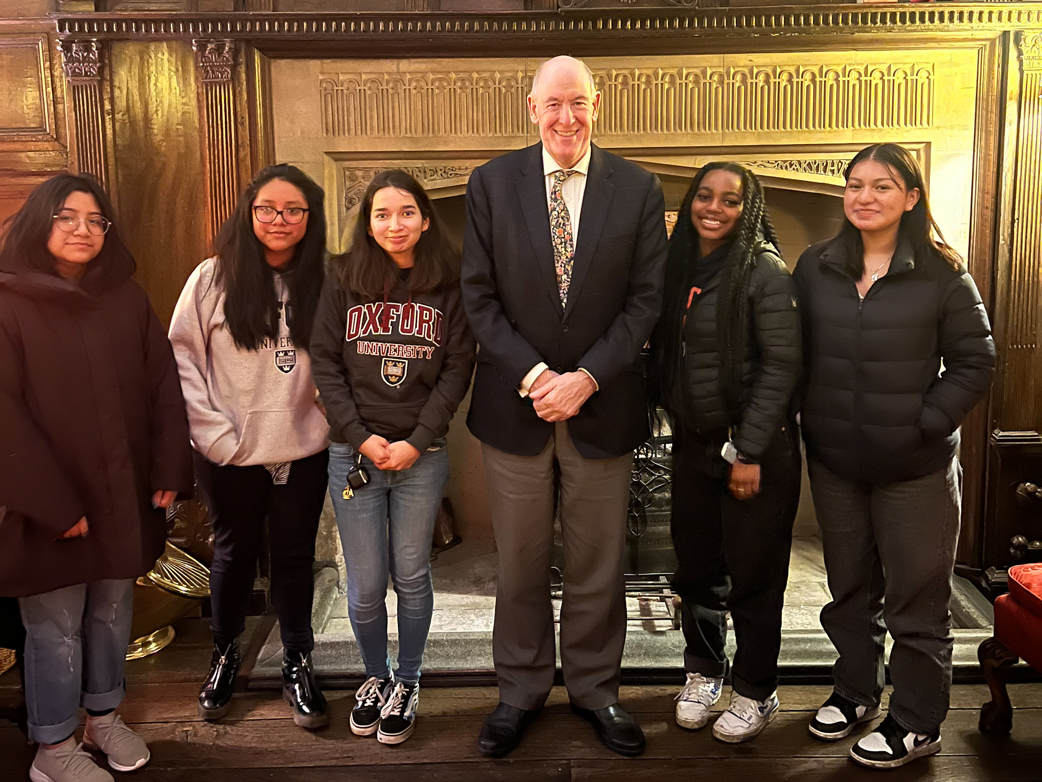 The Warden with five students standing in front of an old fireplace