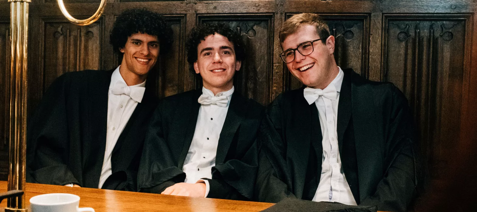 Three smiling students sat in sub-fusc in our dining hall 