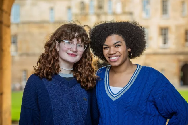 Two female students, one white and one black, stand smiling in front of the Great Quad