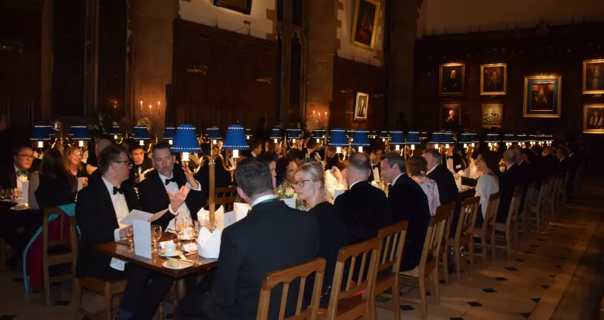 Lots of people sat at long tables in an old dining hall with lots of paintings on the wall at the end