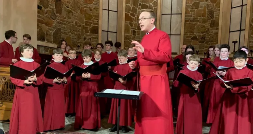 A conductor addresses his audience, with choir behind; all in red robes