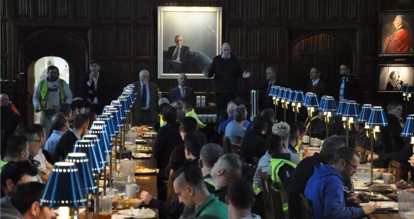 Warden standing on a chair giving a speech to lots of people sat at long tables eating breakfast