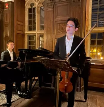 Student holding a violin while another sits behind at a piano