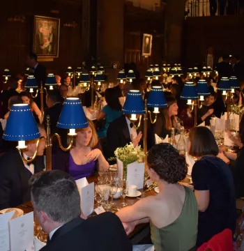 Lots of people sat at long tables in an old dining hall with paintings on the walls