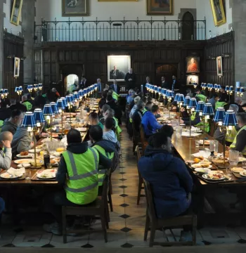 Warden standing on a chair giving a speech to hundreds of people in high vis clothing eating breakfast; all in a medieval Dining Hall