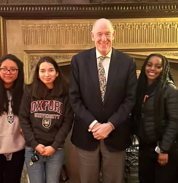 The Warden with five students standing in front of an old fireplace