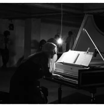B/w image of Jane Chapman perched in front of a harpsichord over which a lightbulb is dangling